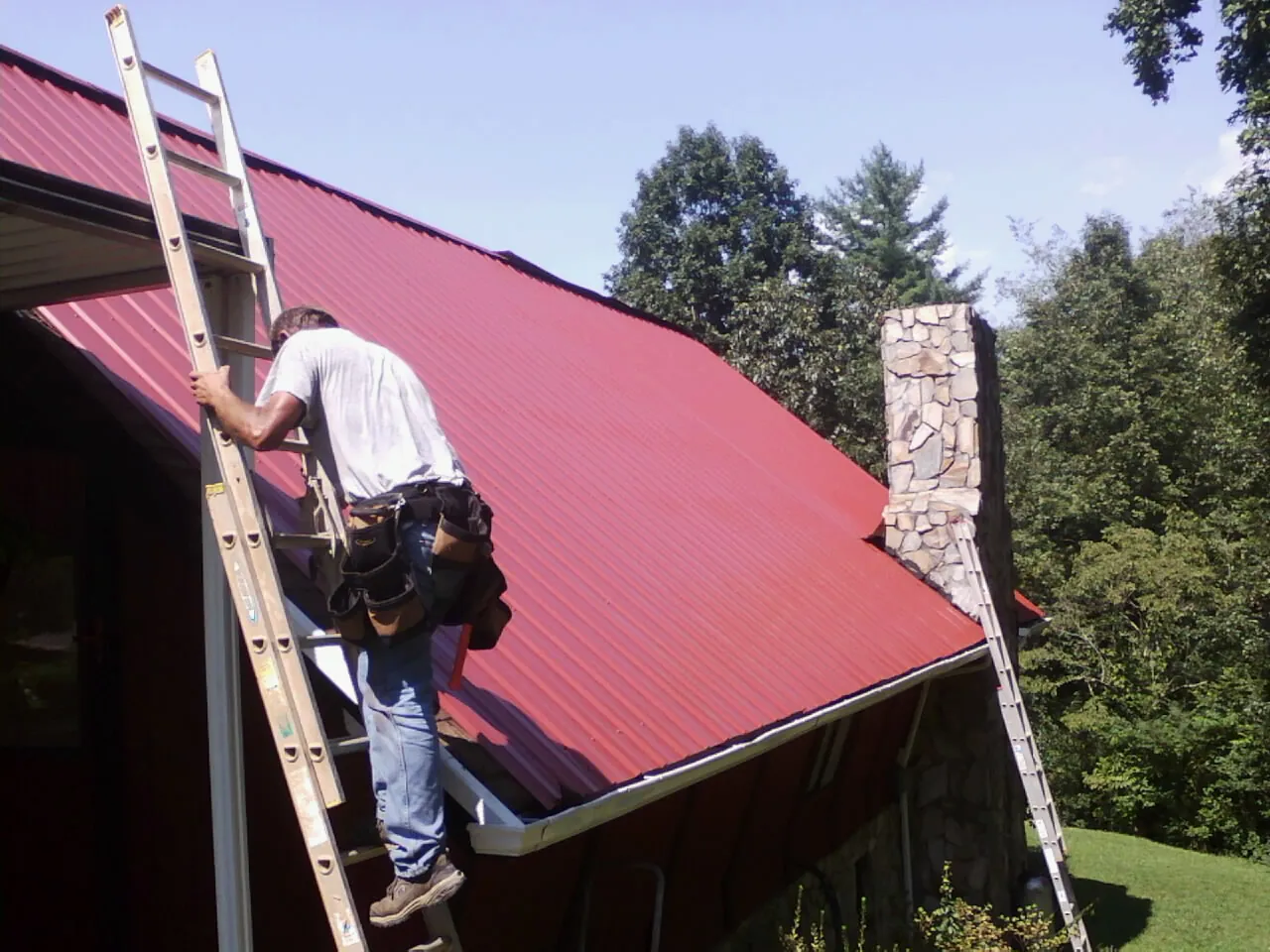 Roofer on a ladder beside a red metal roof and stone chimney — Soffit & Fascia Repair by Roofing Brothers in Oak View